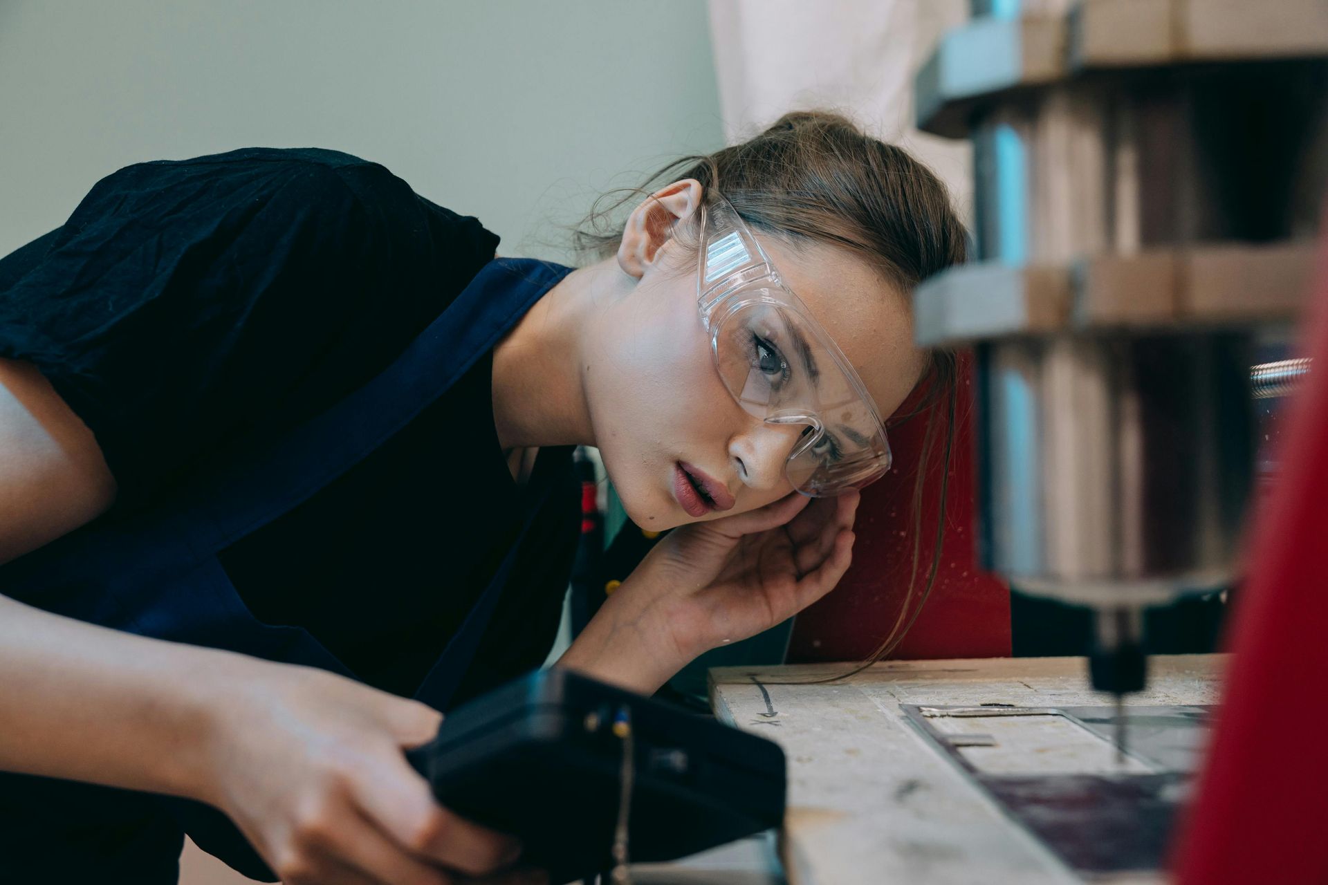 Woman with safety glasses operates CNC machine, focused and leaning in, indoors.