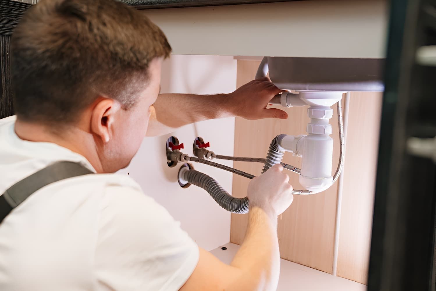Plumber in white shirt working under a sink, adjusting pipes. Inside a light wood cabinet.