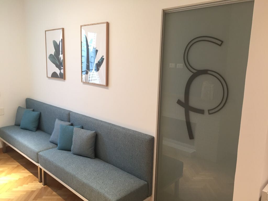 Sala d'attesa con panca blu incorporata, due stampe botaniche incorniciate e una porta in vetro smerigliato con logo.