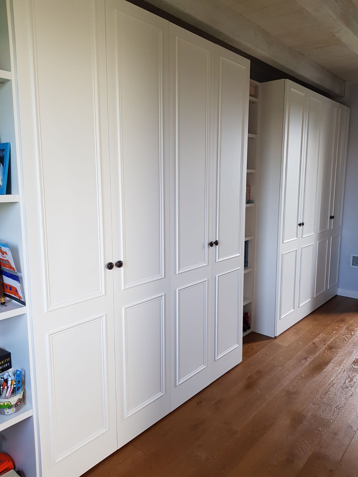 Armadi con pannelli bianchi e pomelli neri sono appoggiati a una parete, accanto a scaffali aperti con oggetti.