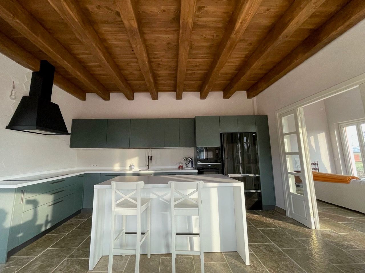 Cucina con mobili color verde acqua, soffitto con travi in ​​legno, isola bianca con sgabelli ed elettrodomestici neri.