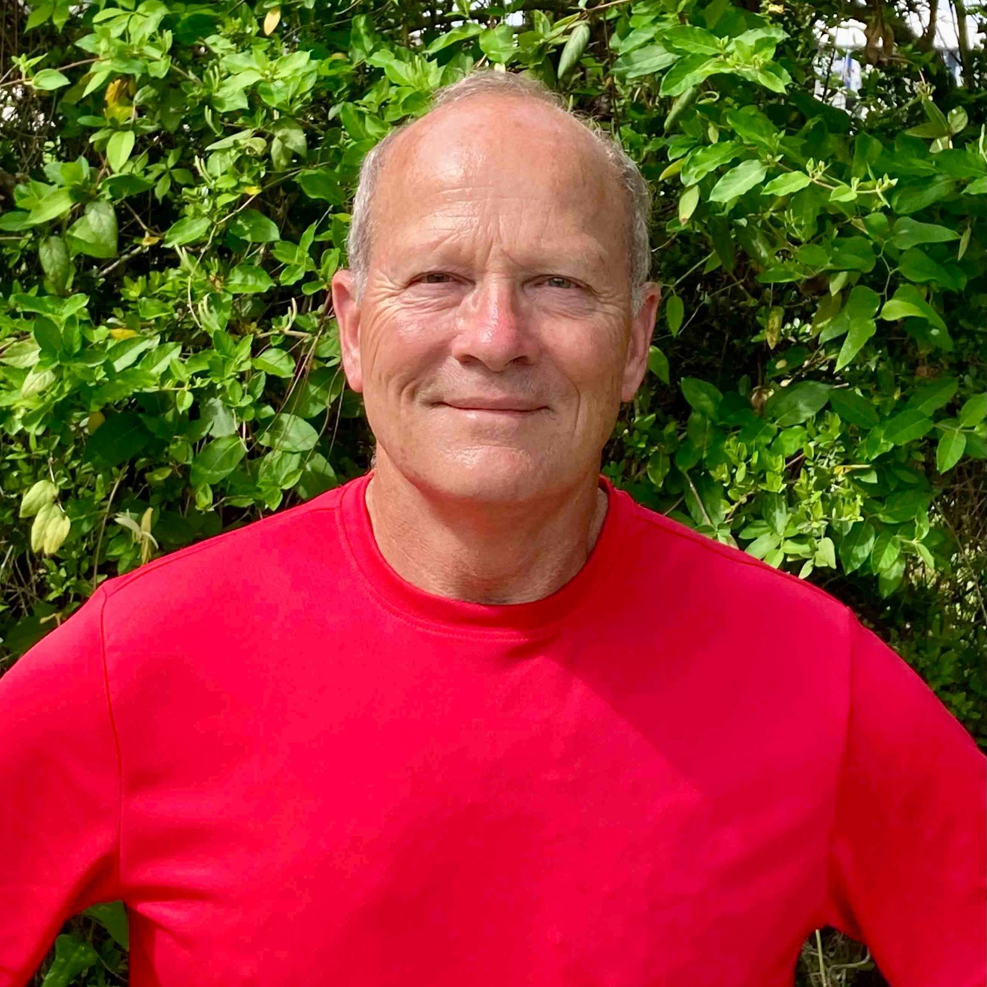 A man in a red shirt is standing in front of a bush.