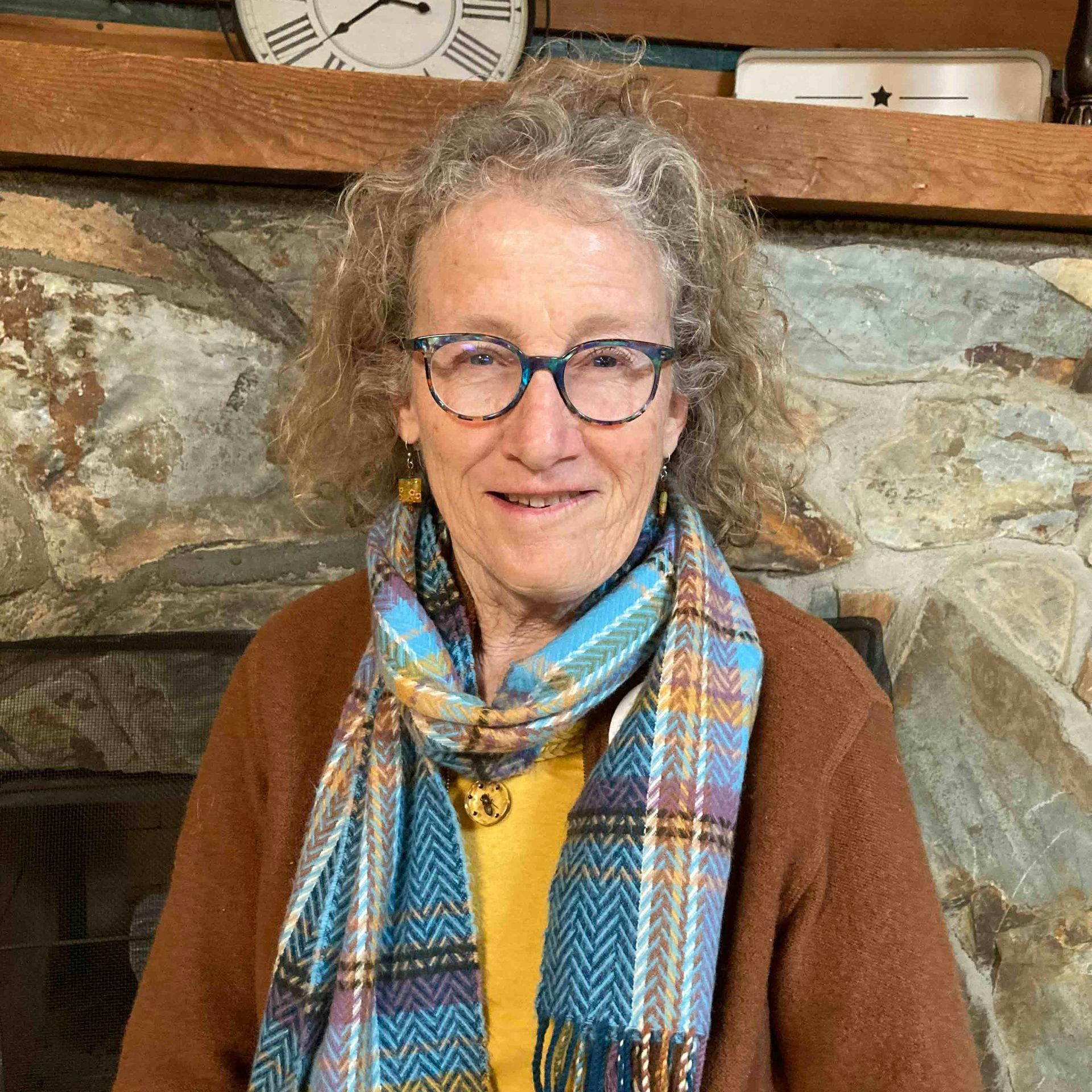 A woman wearing glasses and a scarf is standing in front of a fireplace.