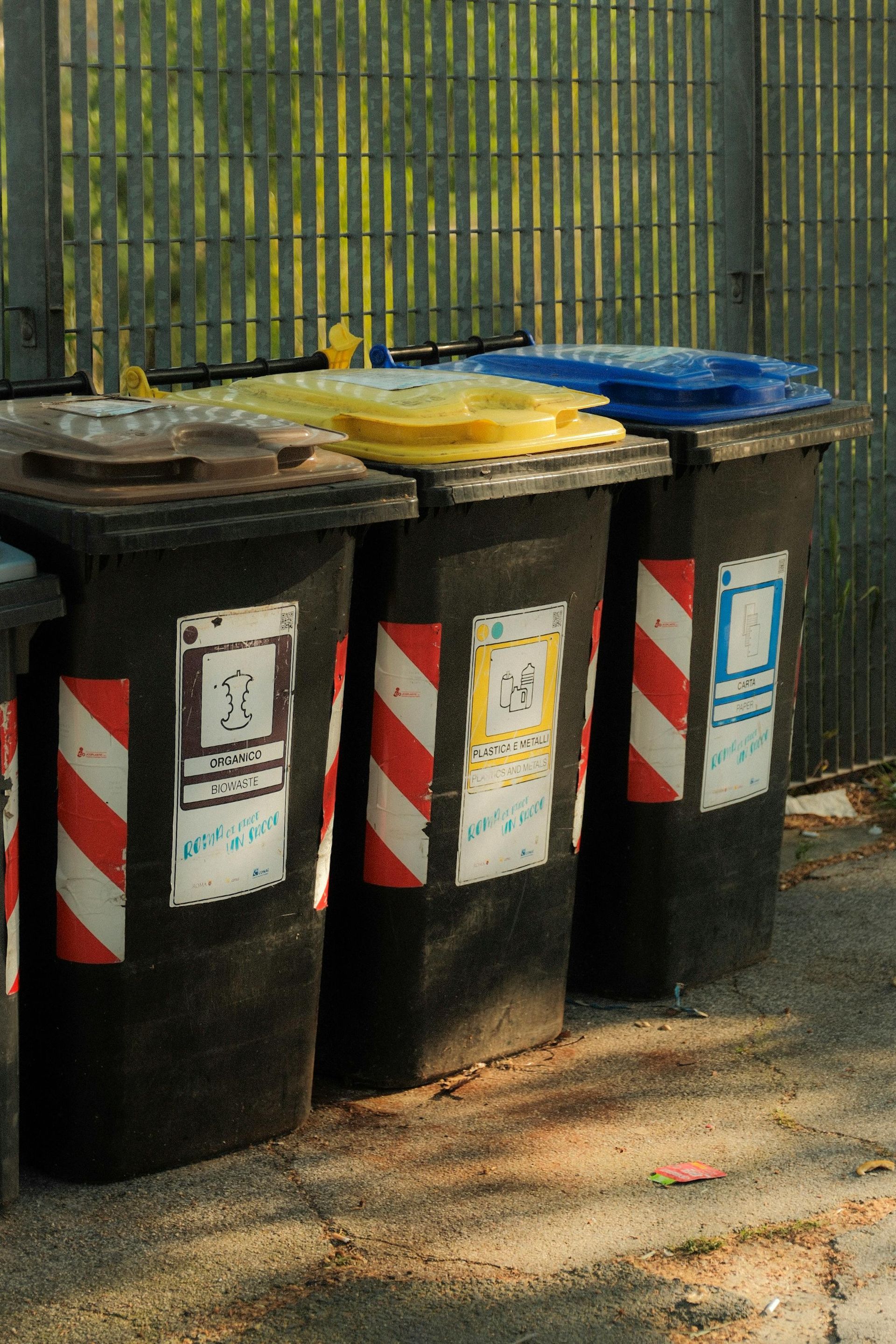 Black recycling bins with yellow and blue lids, red and white striped sides, in front of a metal fence.
