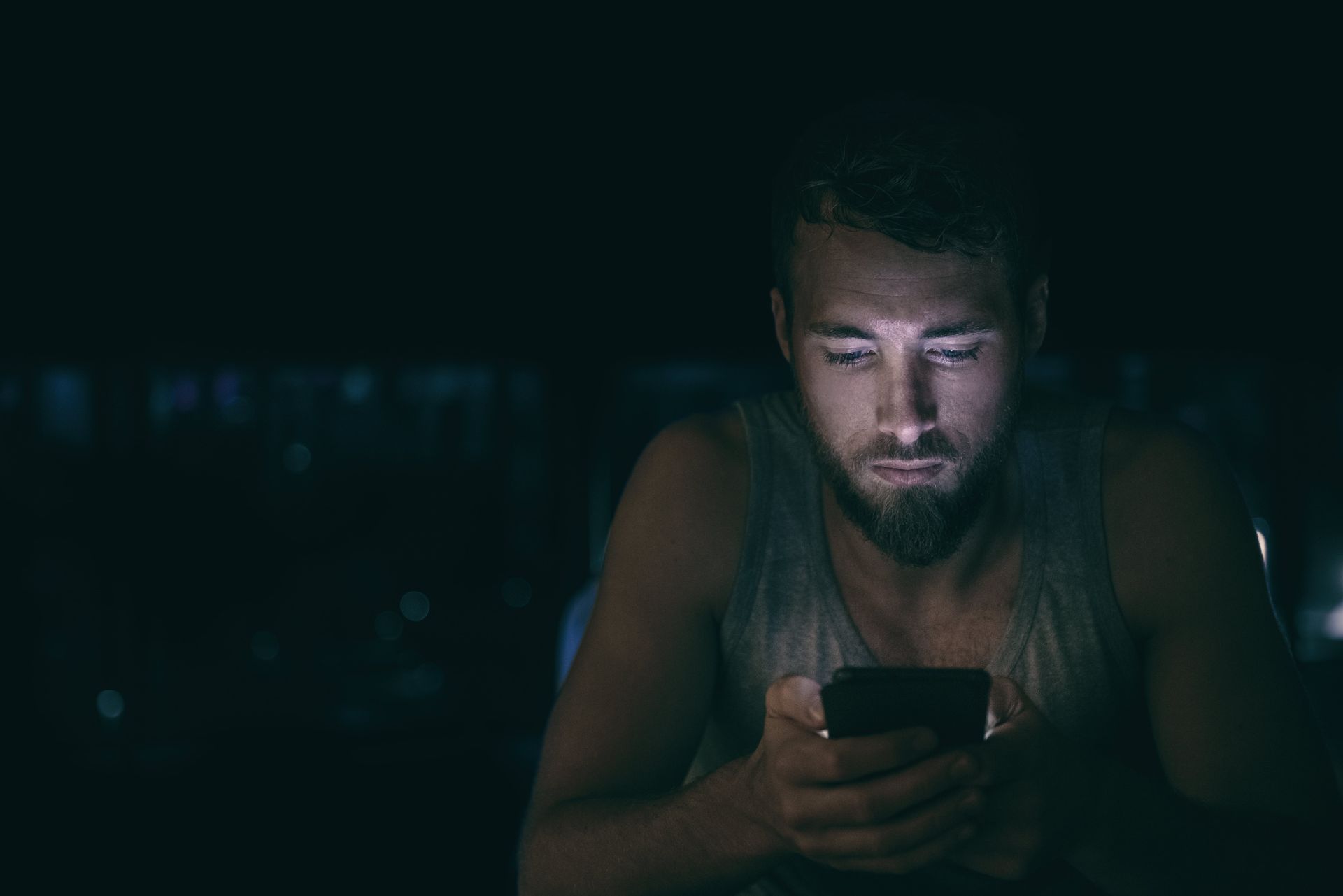 Man in dark, illuminated by phone screen. Gazing intently, with focused expression.