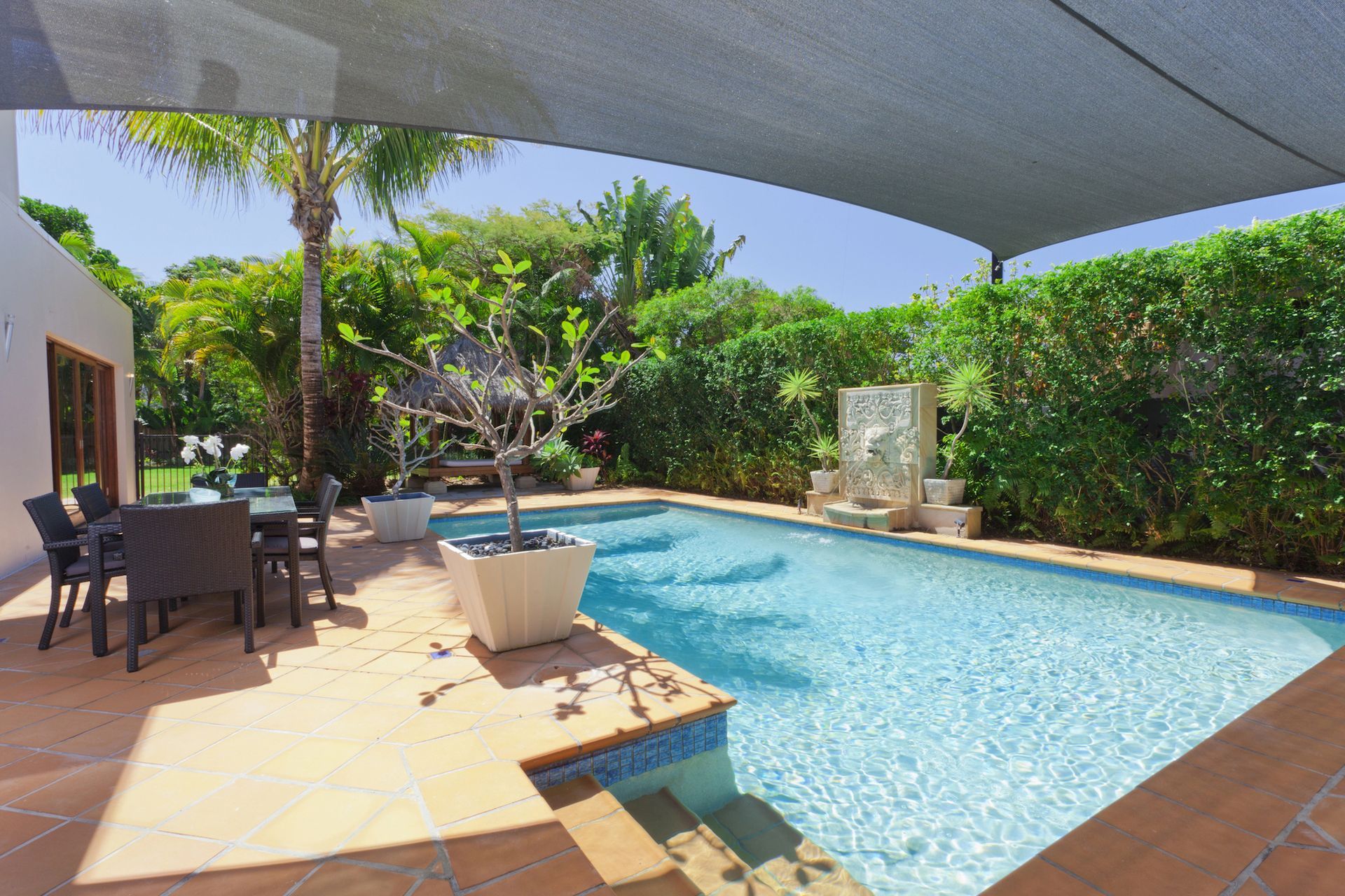 Backyard pool with turquoise water, patio dining set, potted plant, and lush greenery under a shade sail