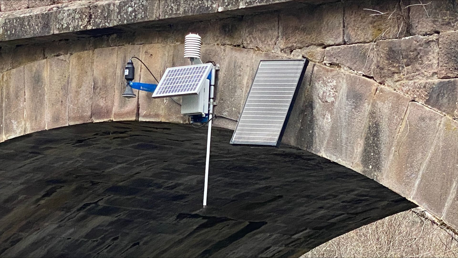 A sensor device attached to the underside of a stone bridge with a solar panel, pole, and antenna.