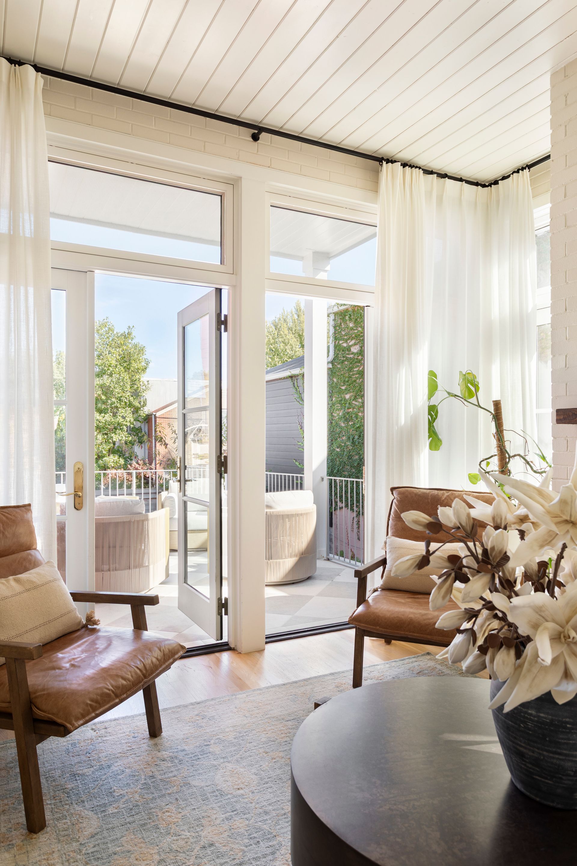 Bright sunlit living room with white curtains, open balcony doors, chairs, and a coffee table with flowers.