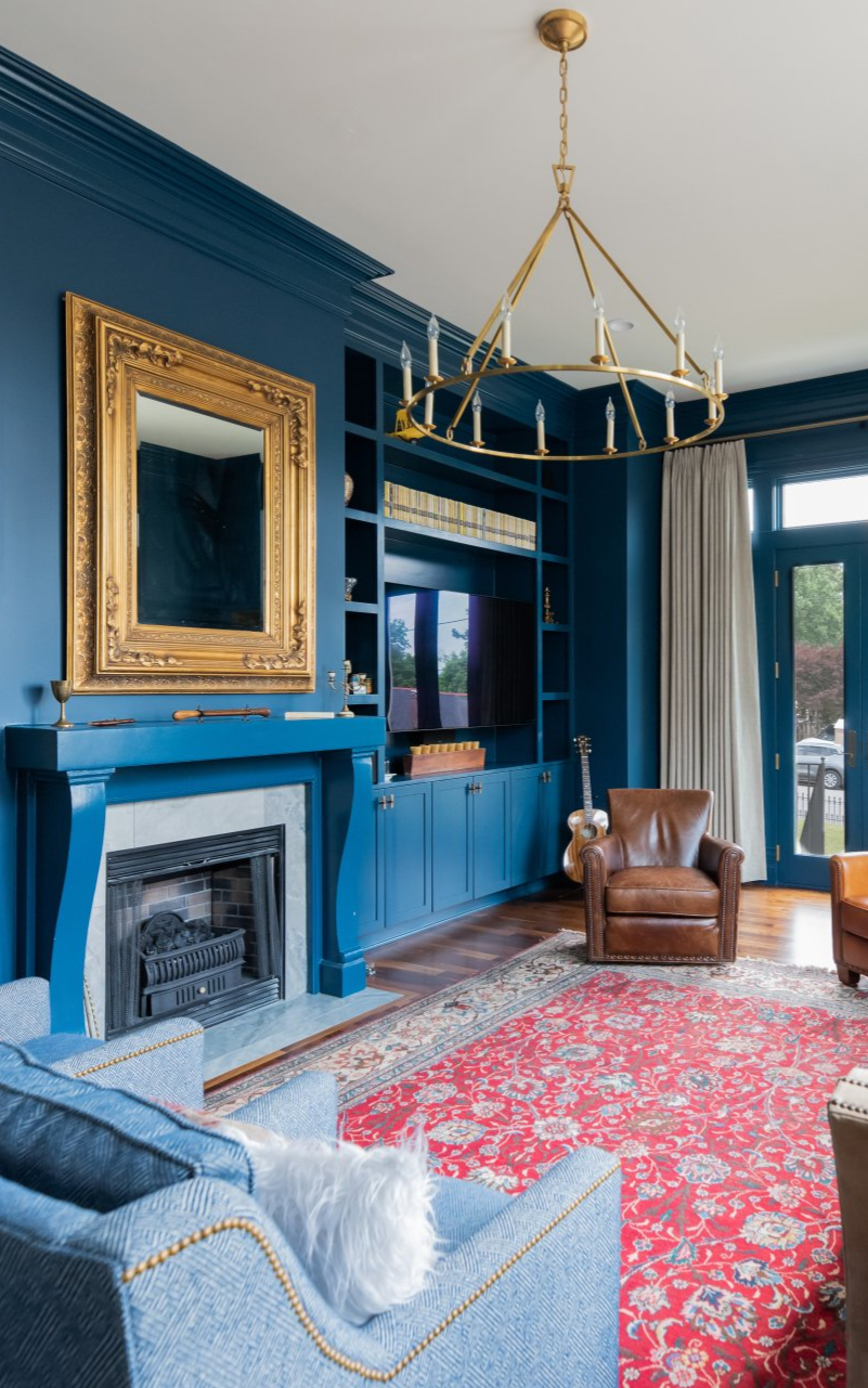 Blue living room with fireplace, gold mirror, red rug, and leather chair by tall windows