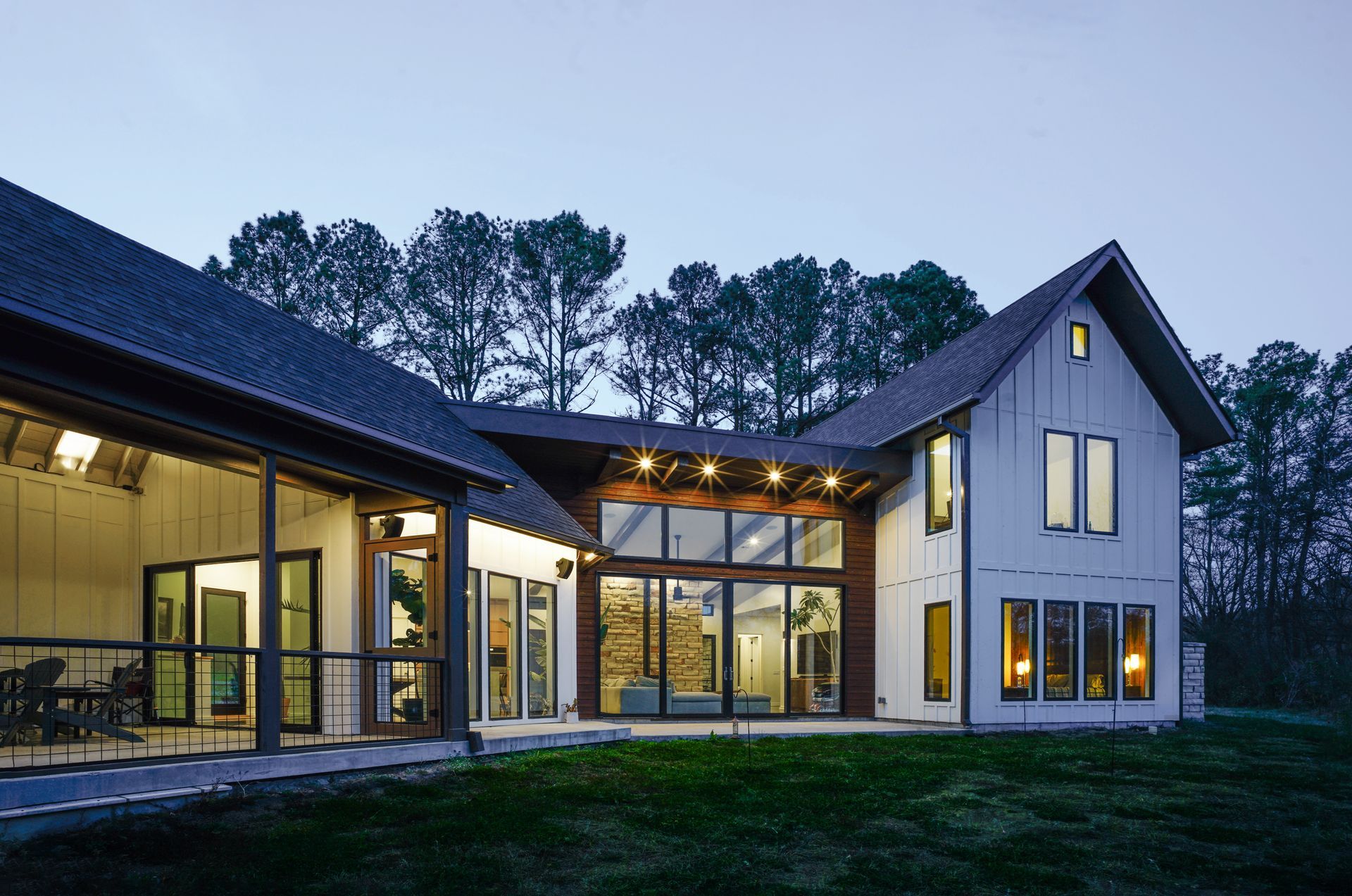 Modern house with large glass windows glowing at dusk, set beside trees and a grassy yard.