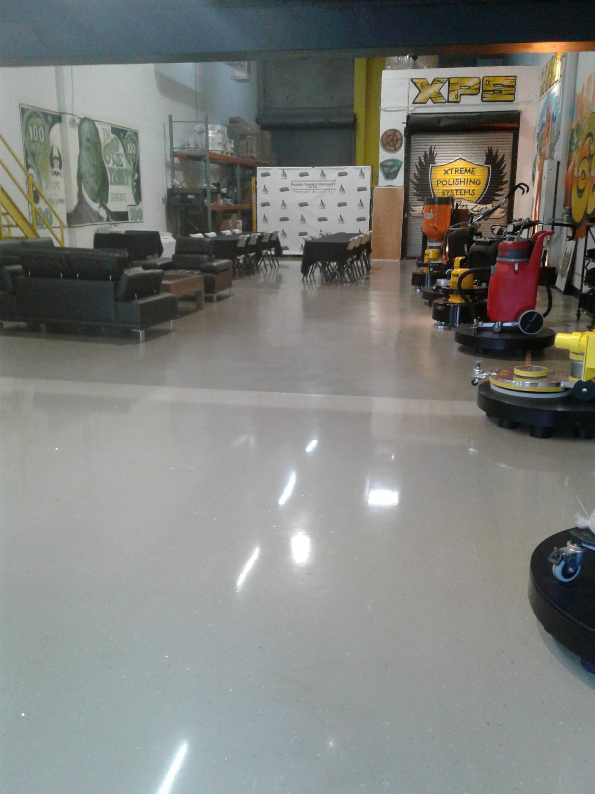 A spacious warehouse with a glossy light-gray floor, black seating, and industrial floor cleaning equipment on the right.