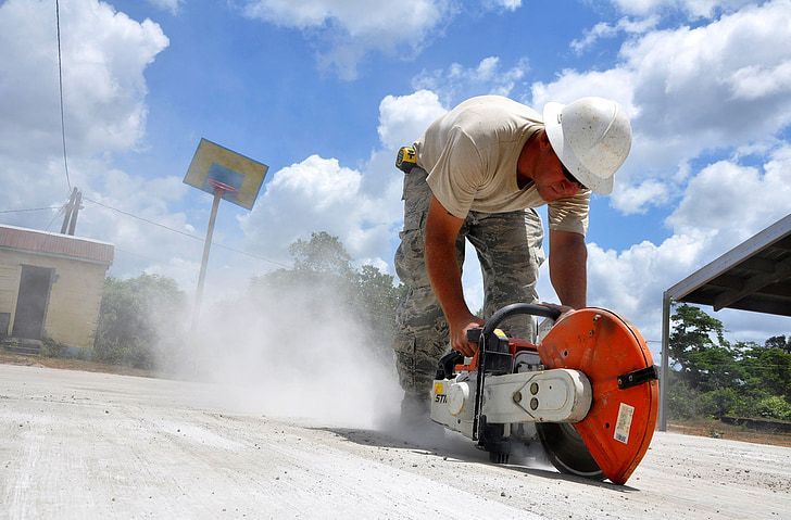 A worker in a hard hat uses a concrete saw, kicking up a large cloud of dust on an outdoor construction site.