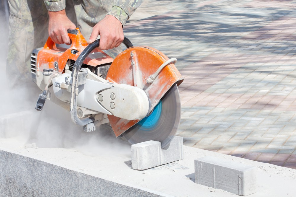 A person uses an orange handheld power saw to cut a concrete block, creating a cloud of dust.