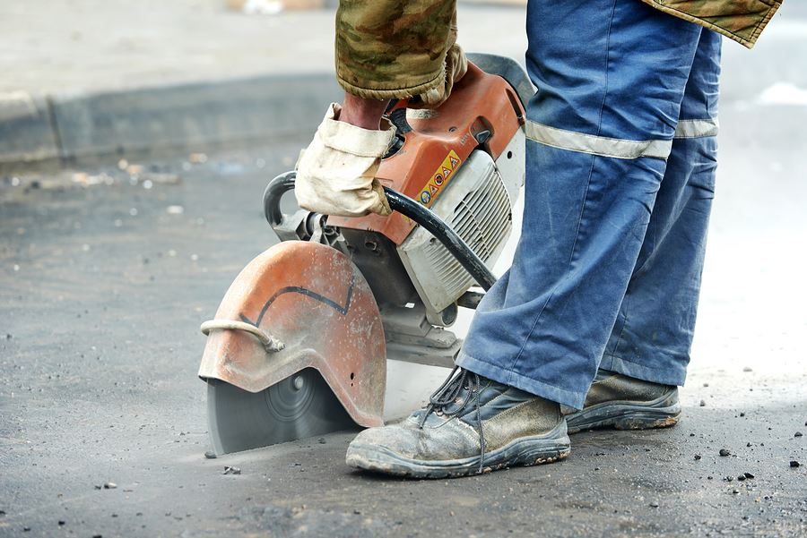 A worker uses a handheld circular concrete saw to cut through a patch of asphalt on a roadway.
