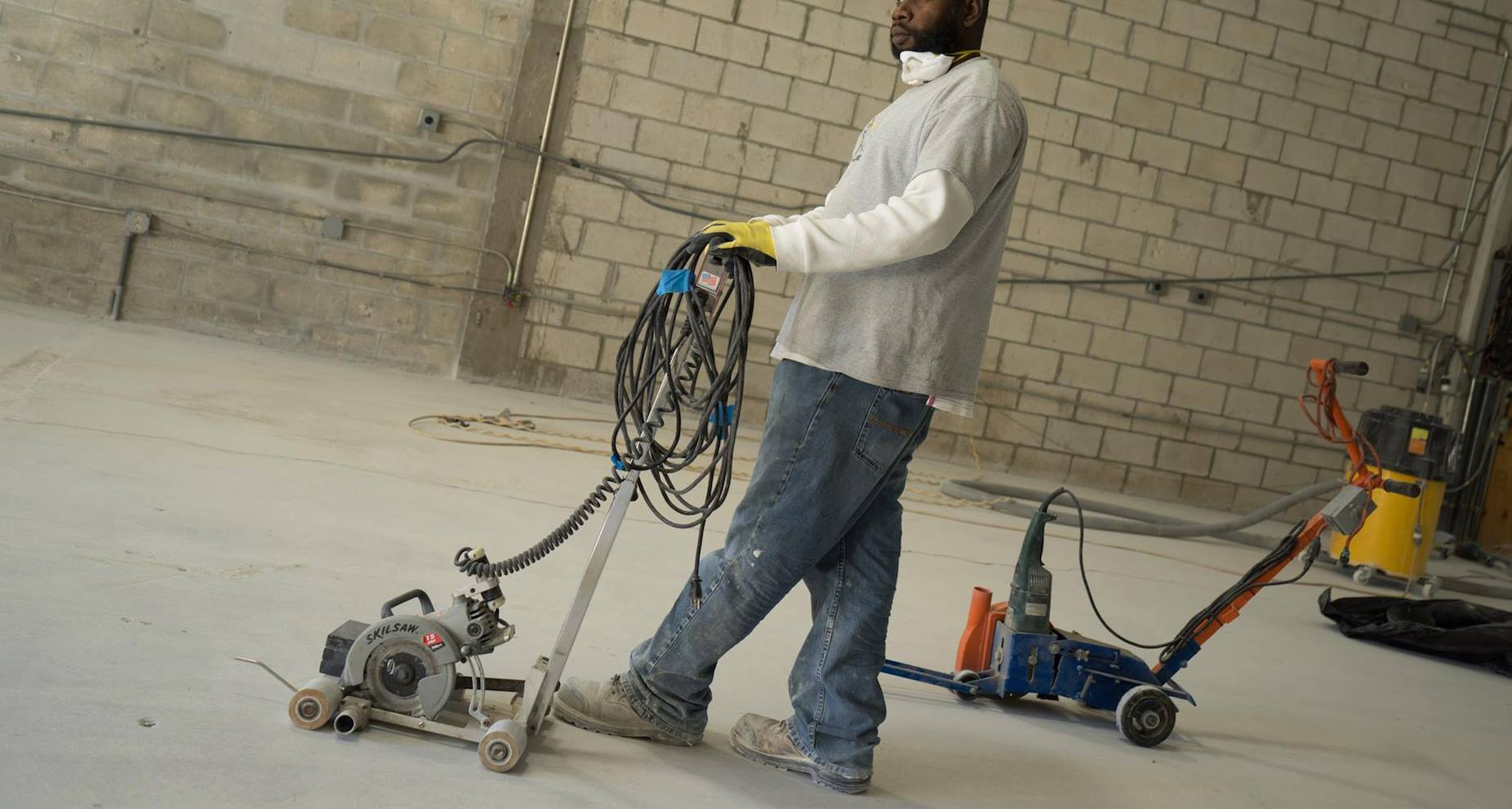 A person operates a floor grinding machine on a concrete surface in an industrial space with unfinished walls.