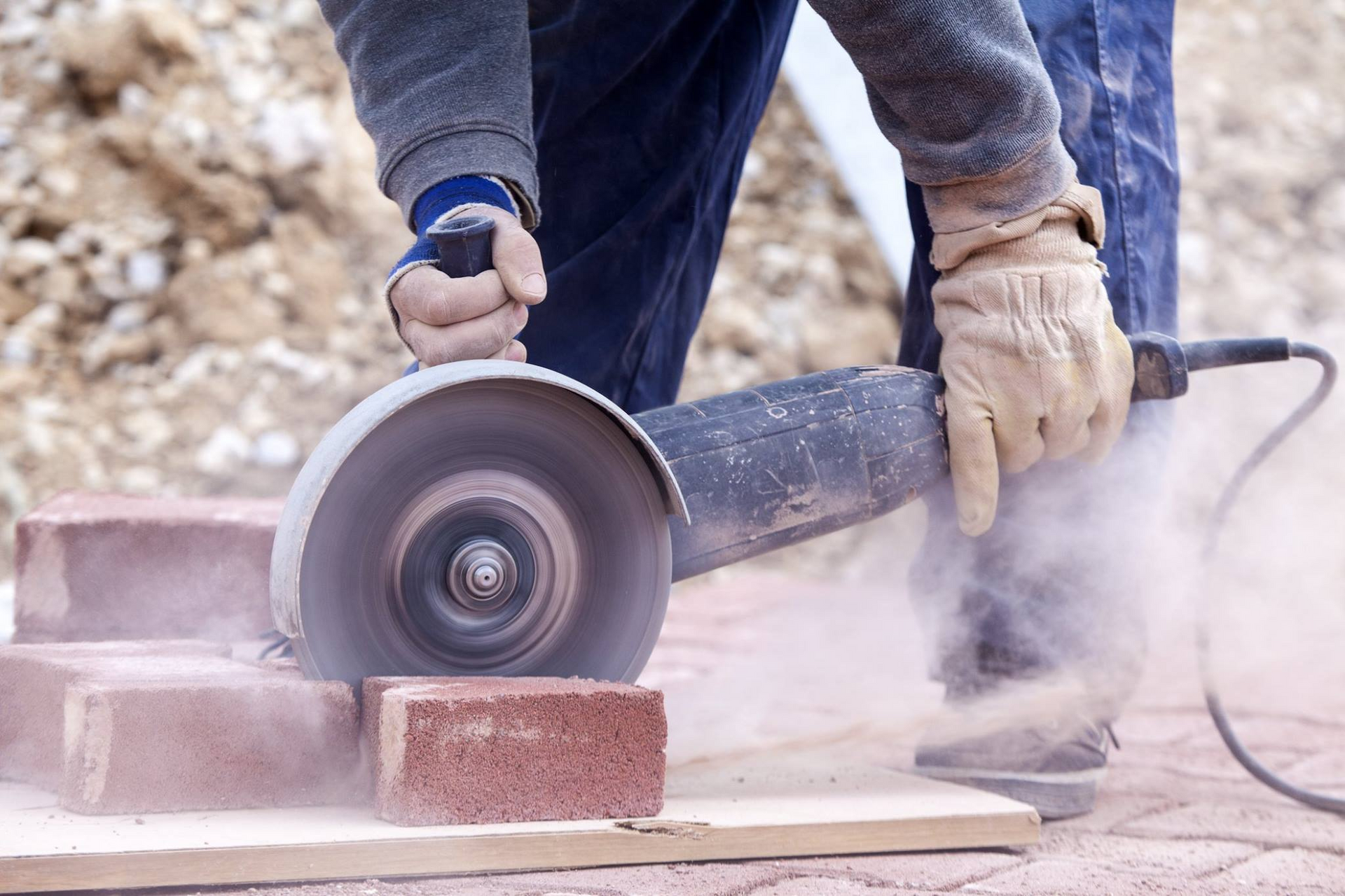 A person wearing work gloves uses an angle grinder to cut through a red brick, creating a cloud of dust.