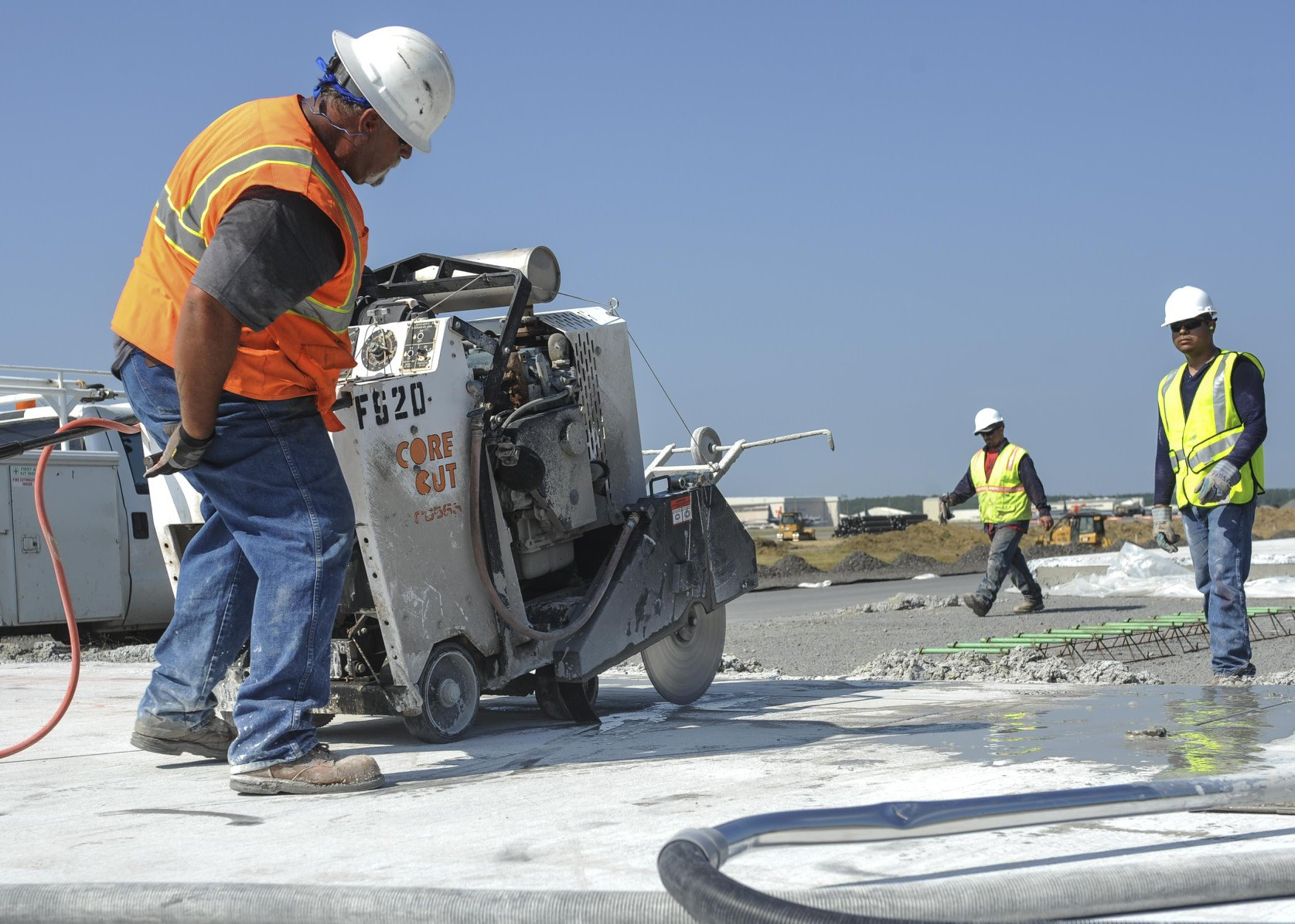 Three construction workers in hard hats and safety vests operate a concrete saw on a sunny job site.