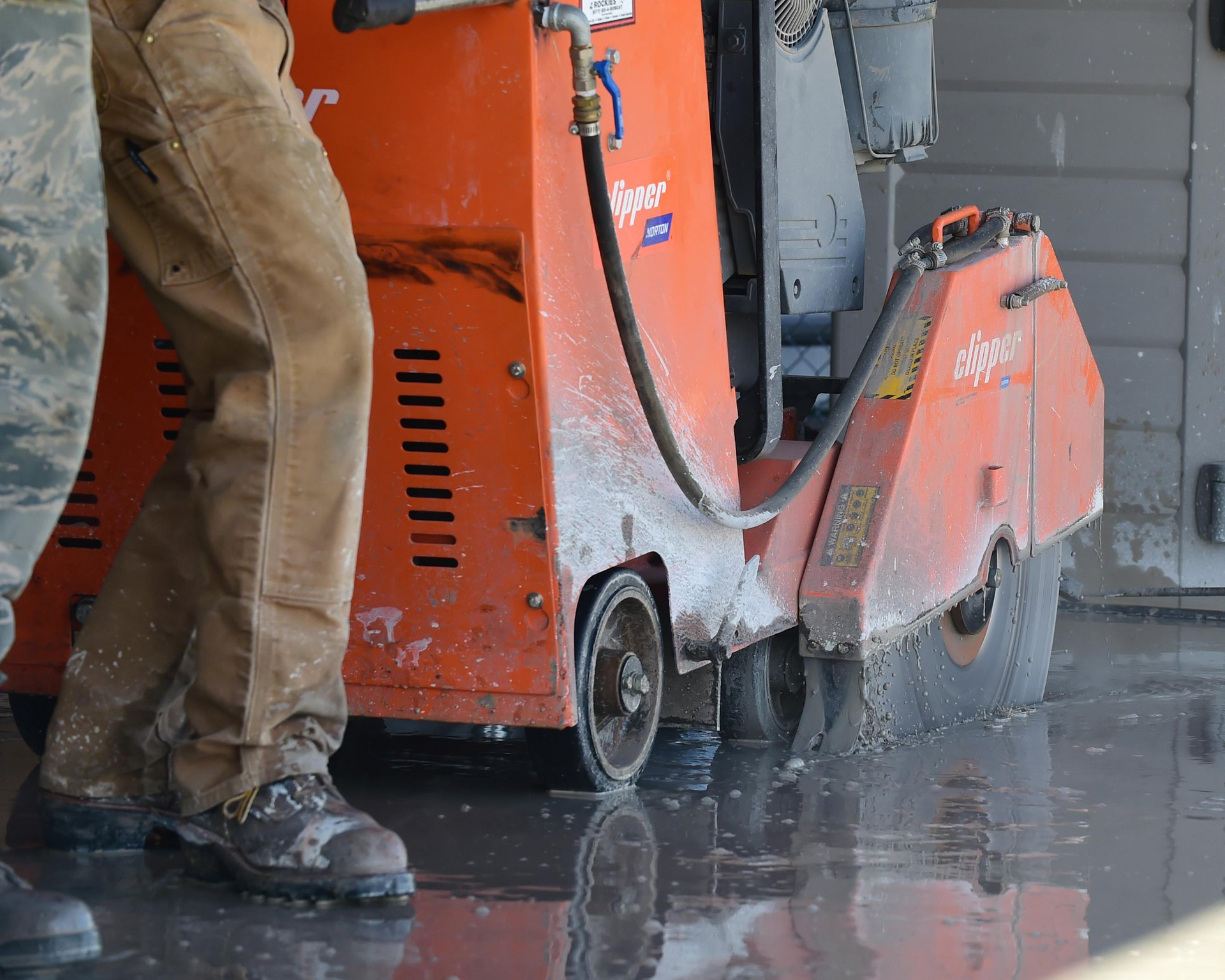 A worker operates an orange floor saw, cutting through a wet concrete surface covered in slurry.