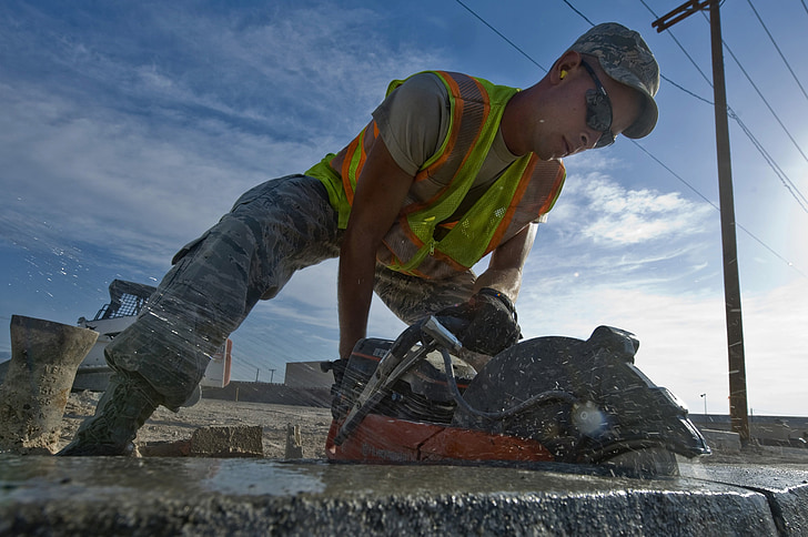 A construction worker in a high-visibility vest uses a handheld power saw to cut into a concrete surface outdoors.