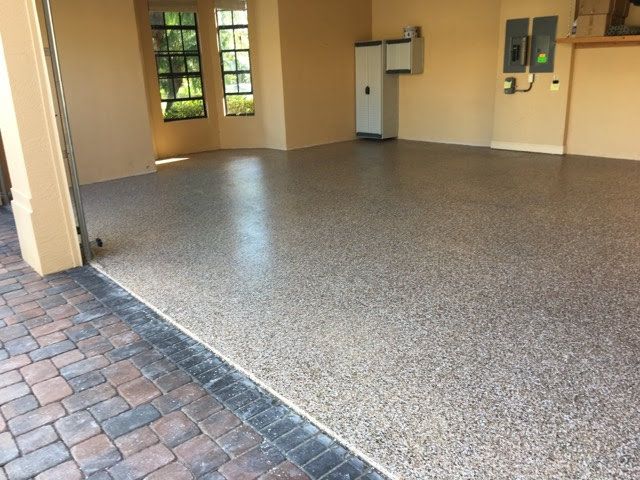 Garage floor featuring a speckled, neutral-toned epoxy finish with an adjacent brick-paver entry and tan walls.
