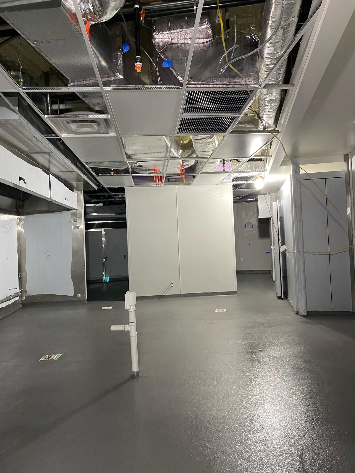An unfinished room with a gray floor, a white partition wall, and exposed industrial ceiling ducts and lighting fixtures.