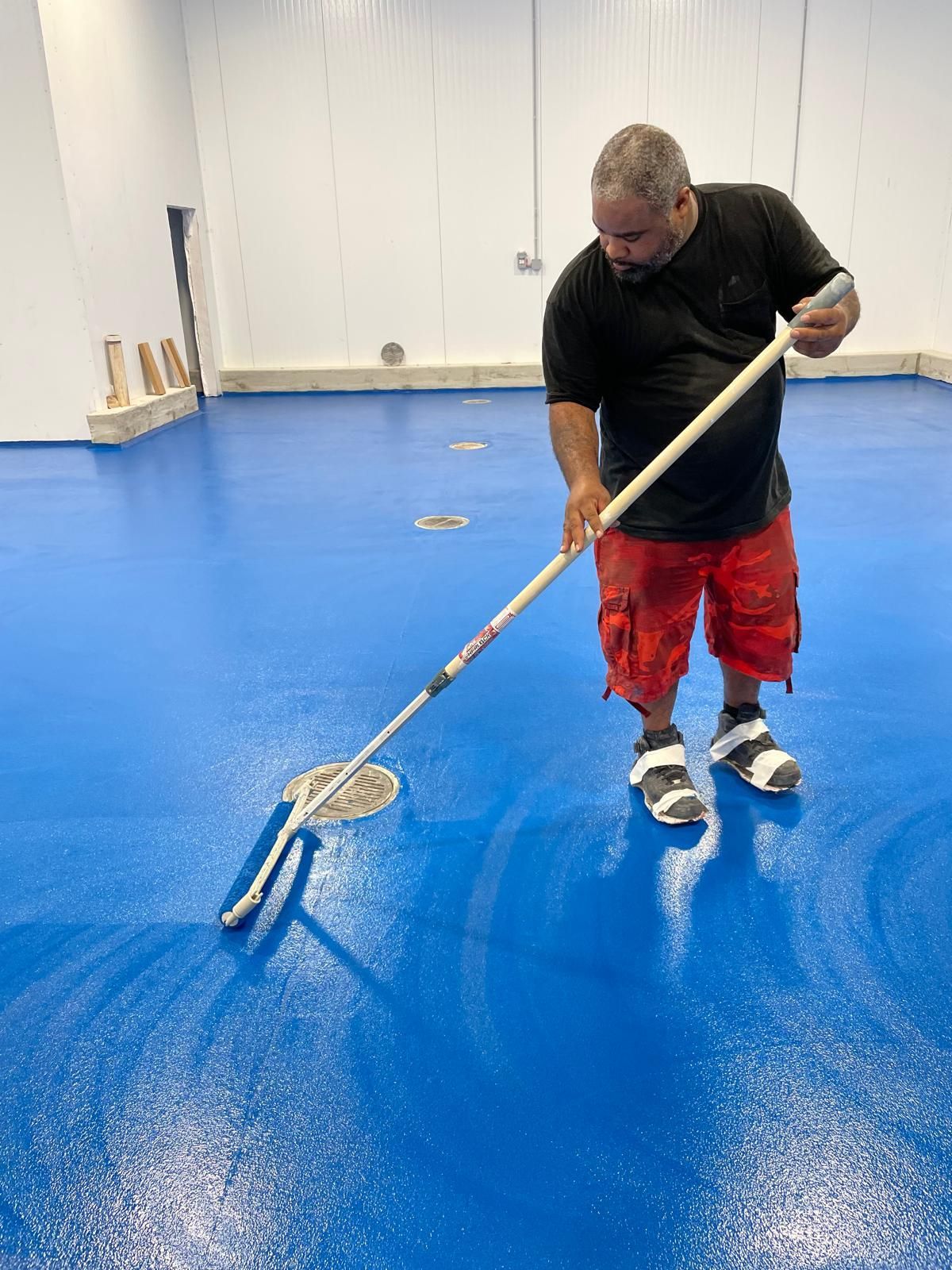 A person in a black shirt and red shorts uses a long-handled roller to apply blue epoxy coating to a warehouse floor.