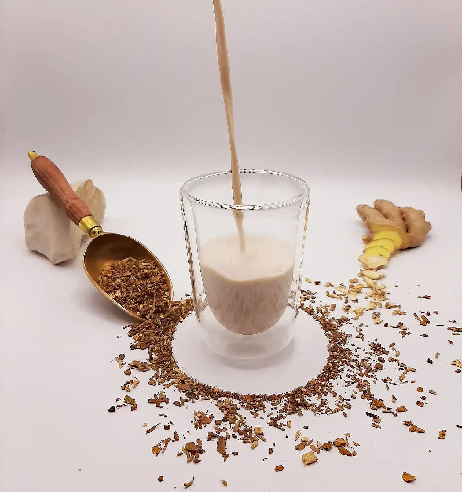 Une boisson est versée dans un verre à double paroi ; des épices et du gingembre entourent le verre sur une surface blanche.