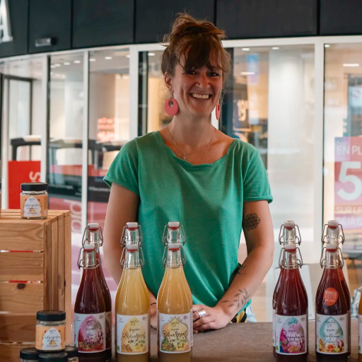 Une femme sourit derrière des bouteilles de boissons, des bocaux et un présentoir en bois dans un magasin.