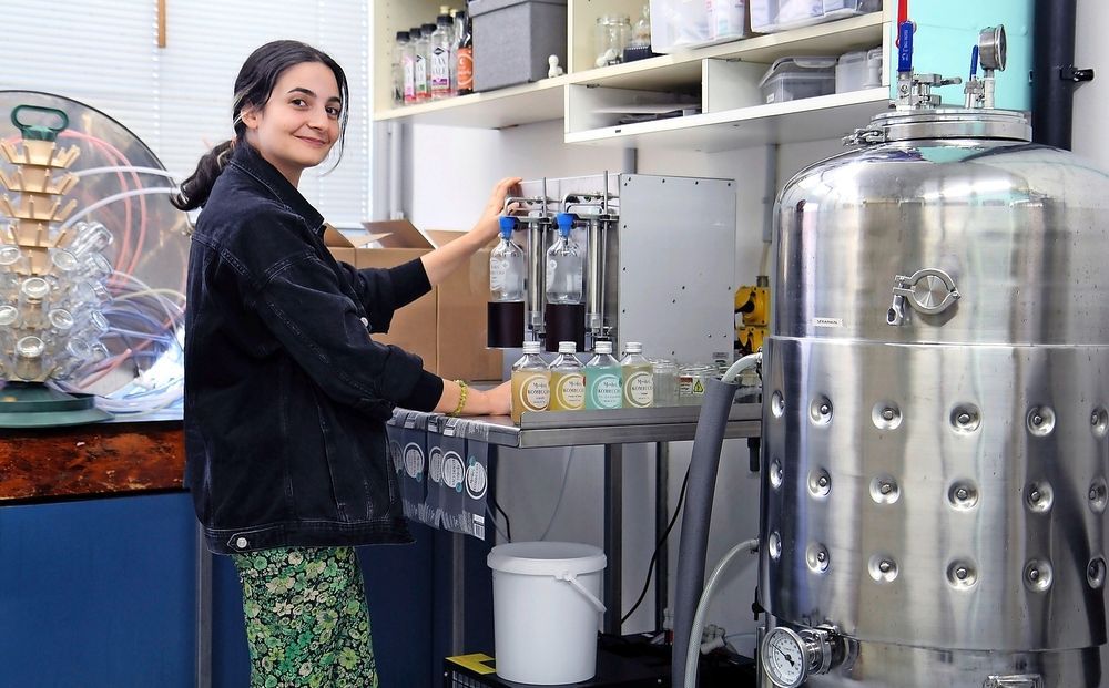 Une femme en blouse blanche remplit des flacons avec un liquide provenant d'une machine. On aperçoit la cuve en acier inoxydable.