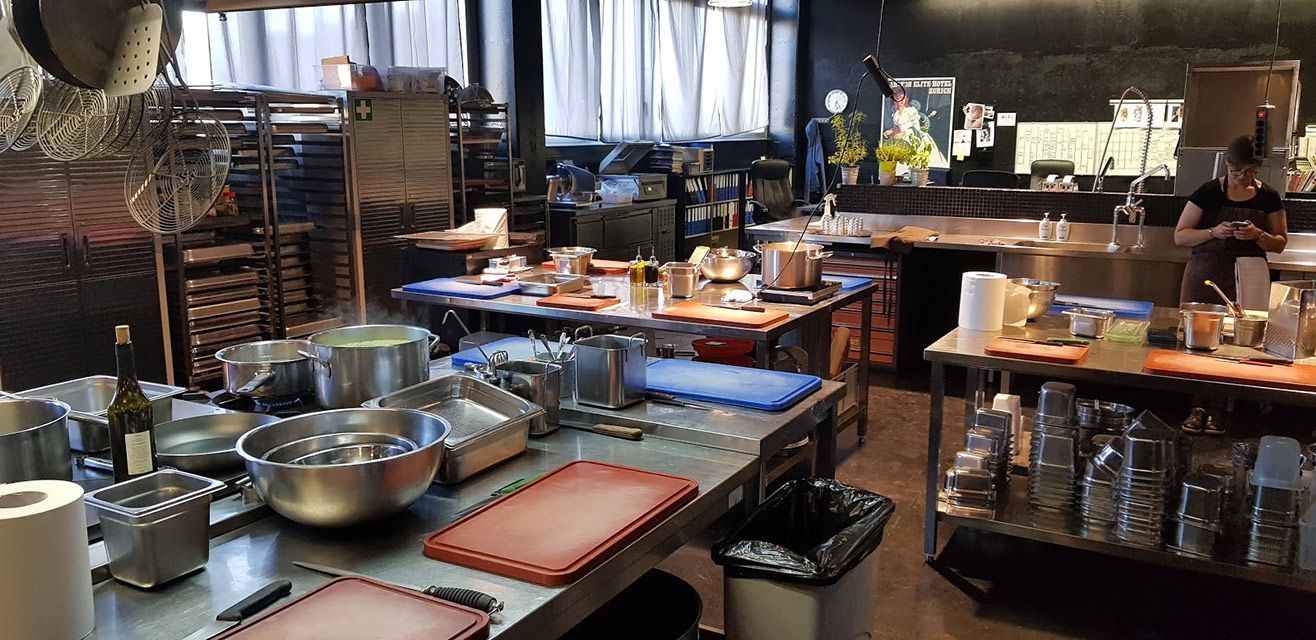 Une cuisine professionnelle animée, avec des comptoirs en acier inoxydable, divers ustensiles de cuisine et une personne qui travaille en arrière-plan.