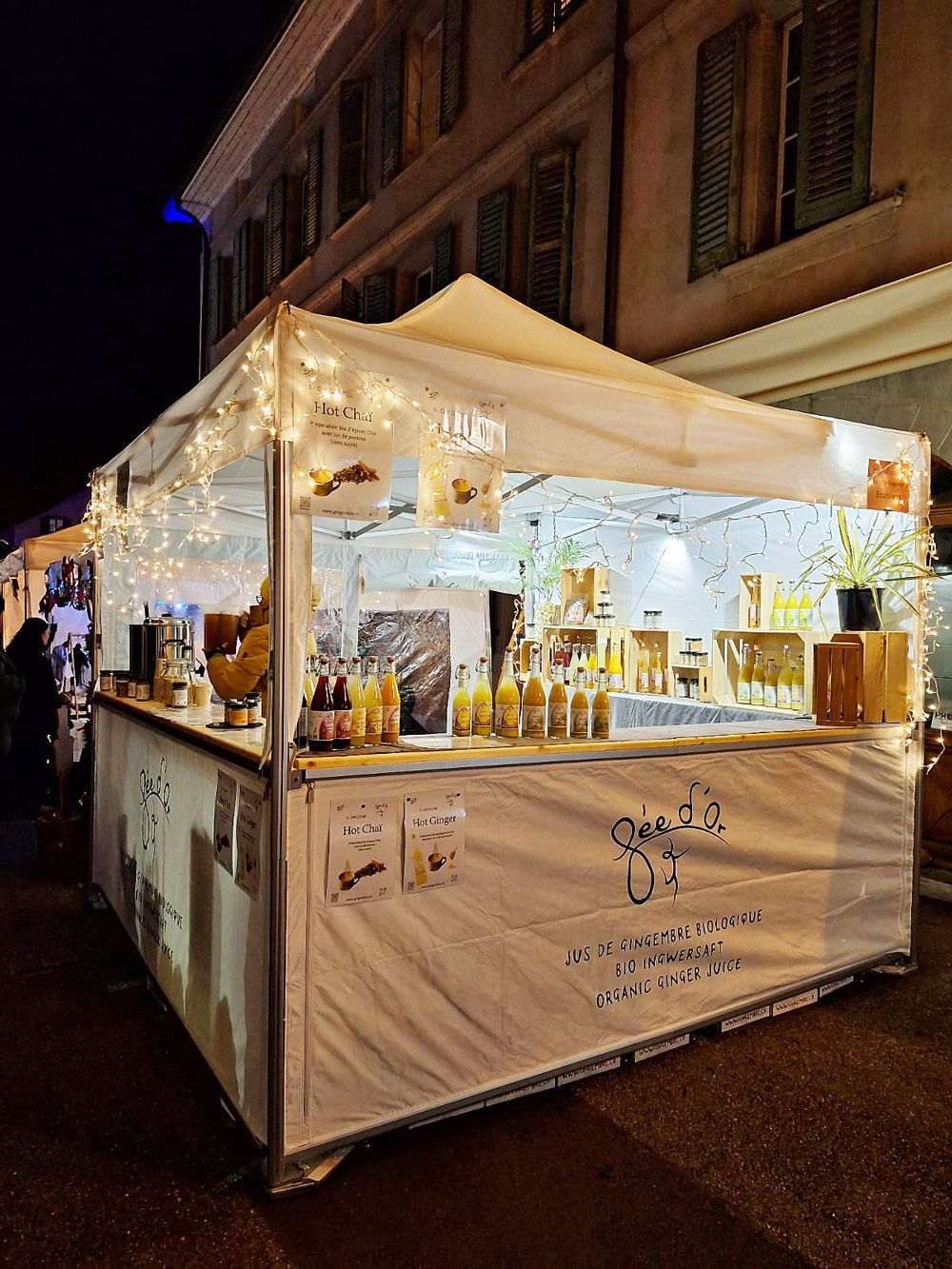 Un étal de vendeur illuminé la nuit, proposant des bouteilles et des bocaux ornés d'une banderole.