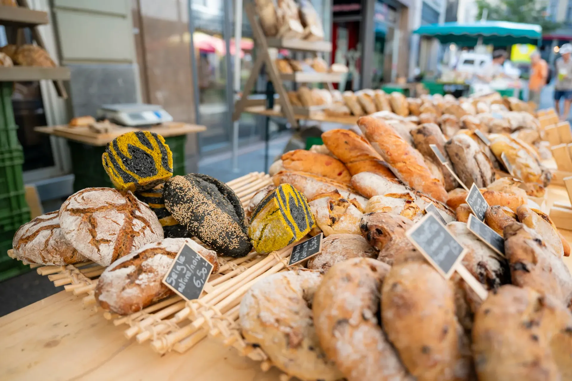 Des pains de formes et de couleurs variées sont présentés sur une table en bois, sur un marché en plein air.