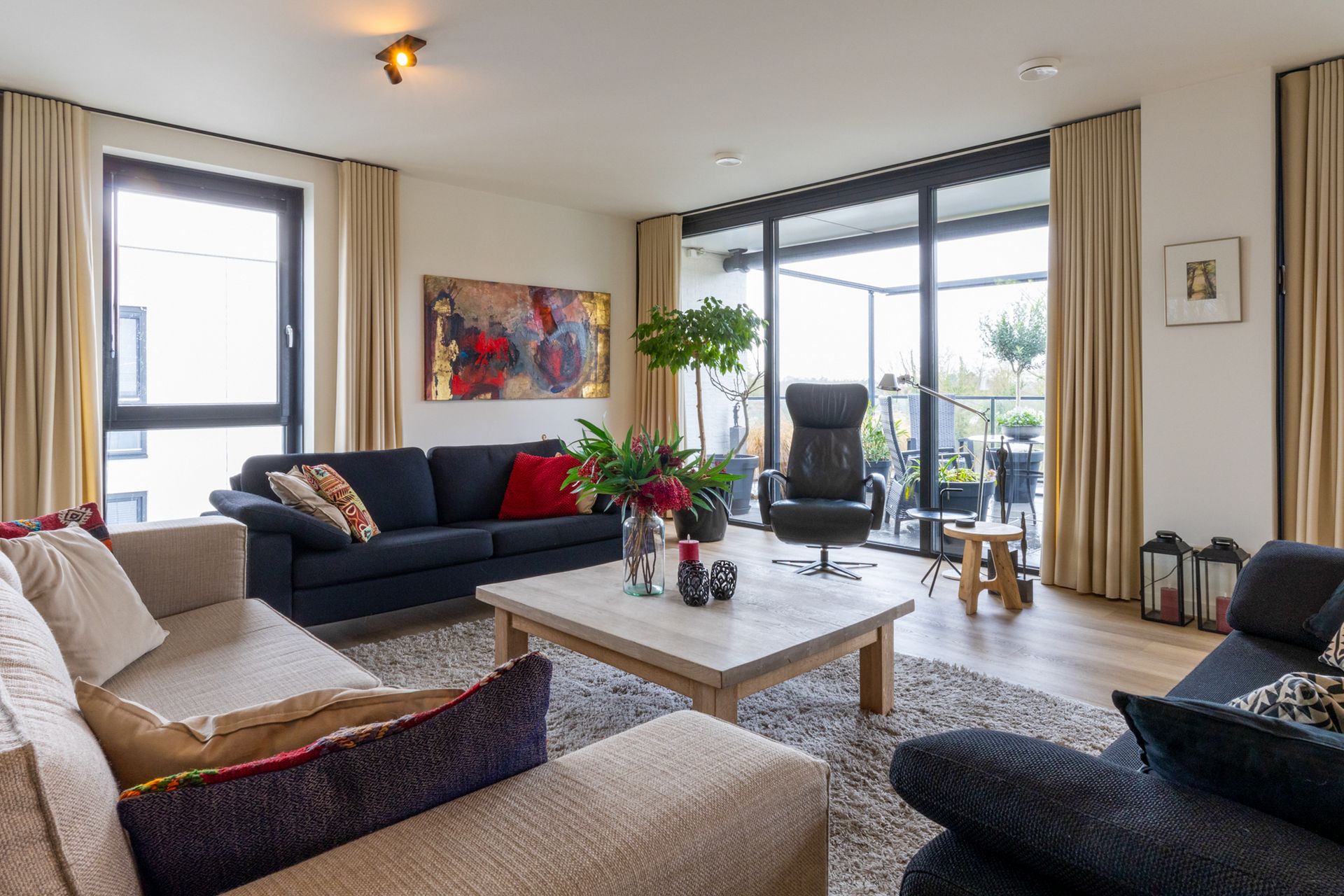 Een moderne woonkamer met maar liefst ruimte voor 3 royale banken. In beige en zwart, en een zwarte sofa, een houten salontafel, een hoogpolig vloerkleed en grote ramen van vloer tot plafond, die toegang geven tot het zeer grote balkon.