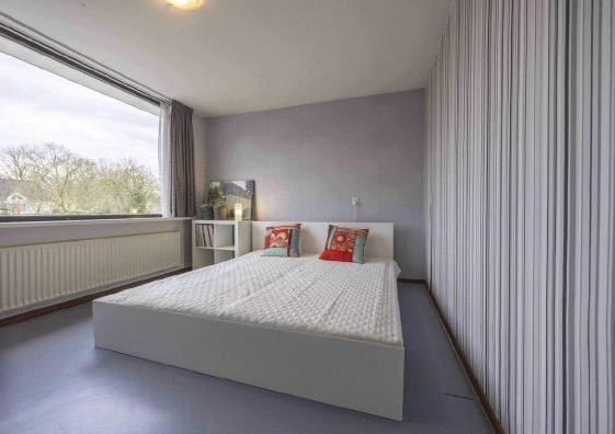Na restyling, een moderne, minimalistische slaapkamer met een wit bed, grijze muren, een groot raam met uitzicht de tuin. De gekleurde kussen geven sfeer aan deze kamer.