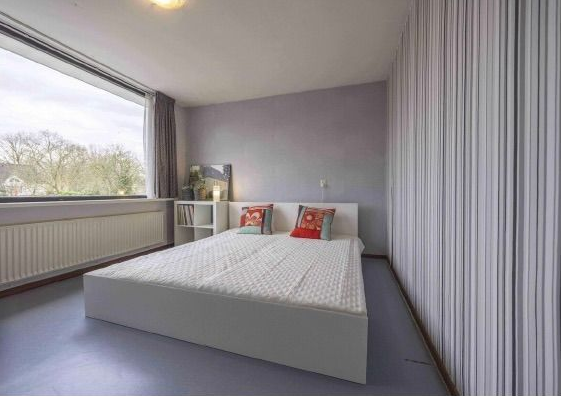 Na restyling, een moderne, minimalistische slaapkamer met een wit bed, grijze muren, een groot raam met uitzicht de tuin. De gekleurde kussen geven sfeer aan deze kamer.