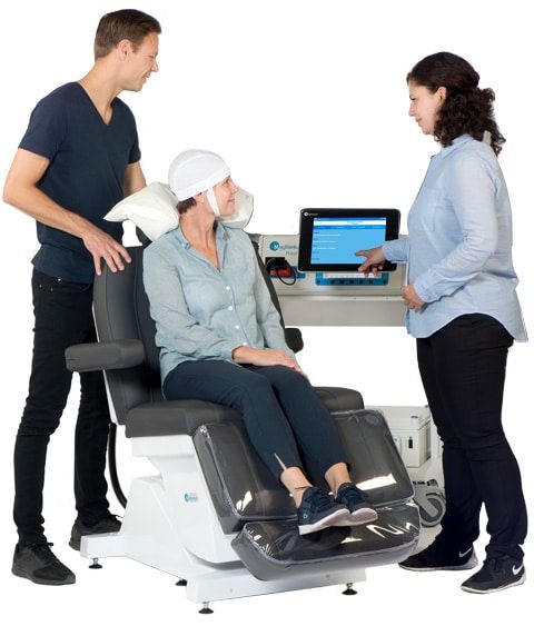 a woman is sitting in a TMS chair with two people standing around her .