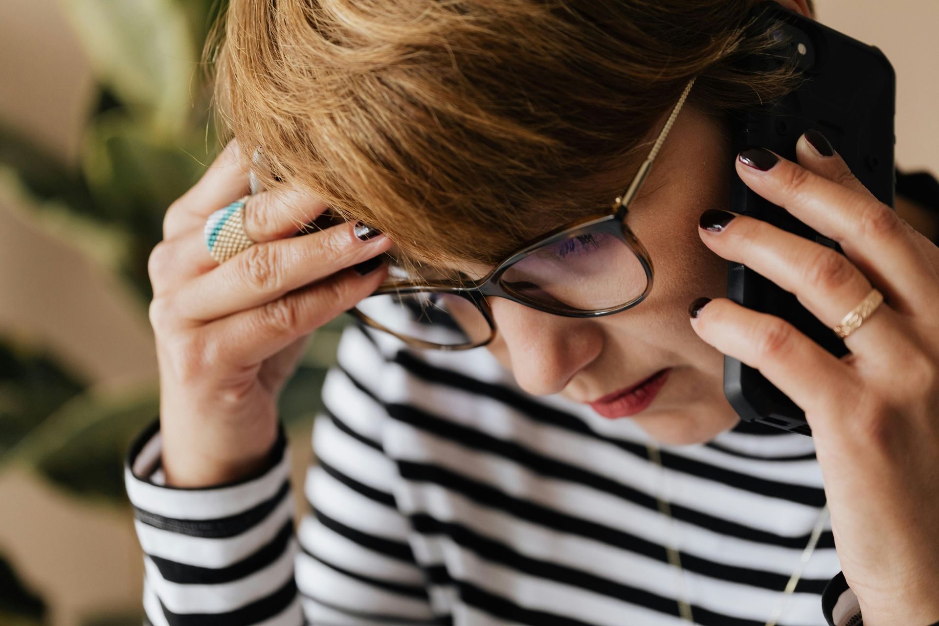 Woman in glasses on the phone, hand to head, striped shirt, concerned expression.