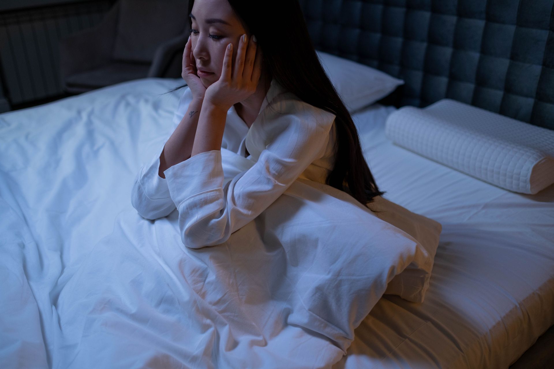 Woman sitting on a bed at night, resting her face in her hands, looking tired or worried.