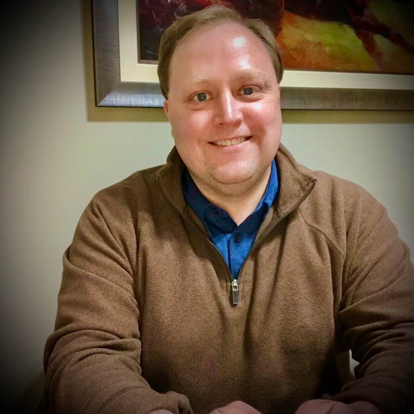 A smiling man in a brown sweater with a blue shirt underneath, seated indoors near artwork.