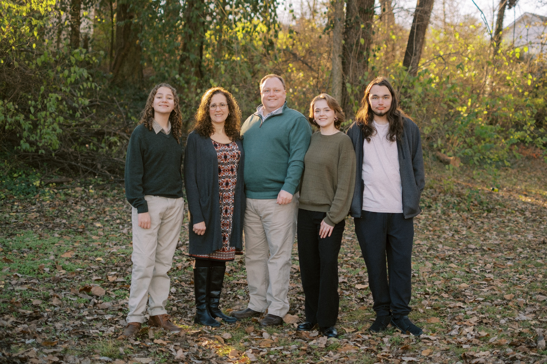 Family of five posing outside in front of trees; smiling, dressed in sweaters and pants.