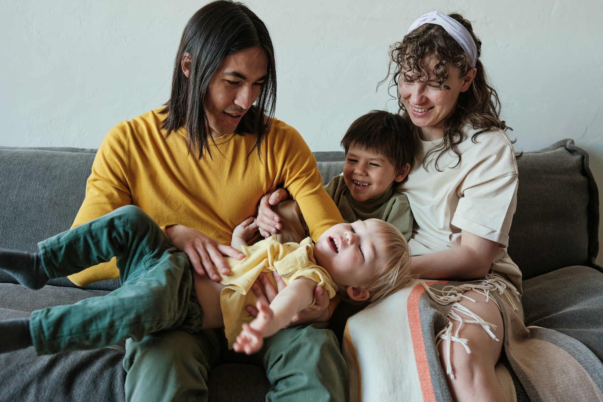 Family of four on a sofa; parents tickle two young children.