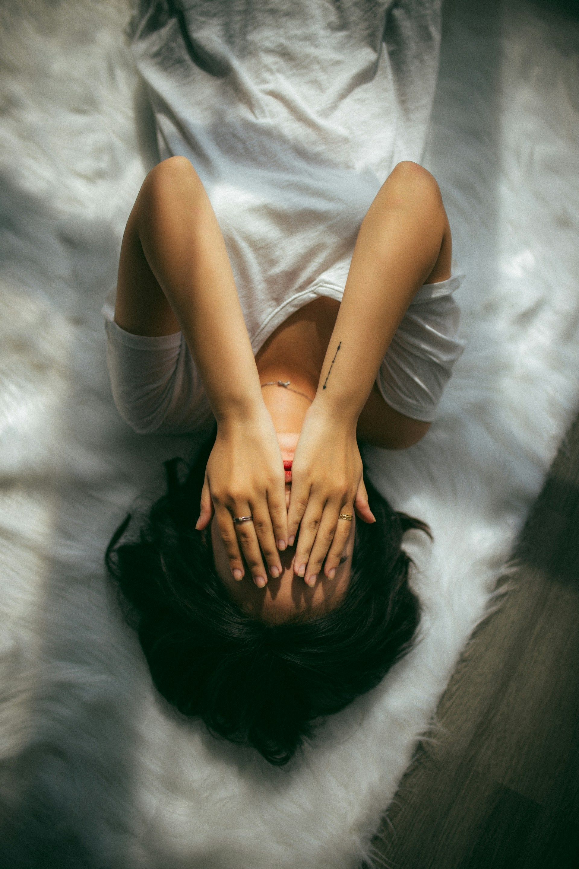 Woman lying on white fur, hands covering her face, dark hair.