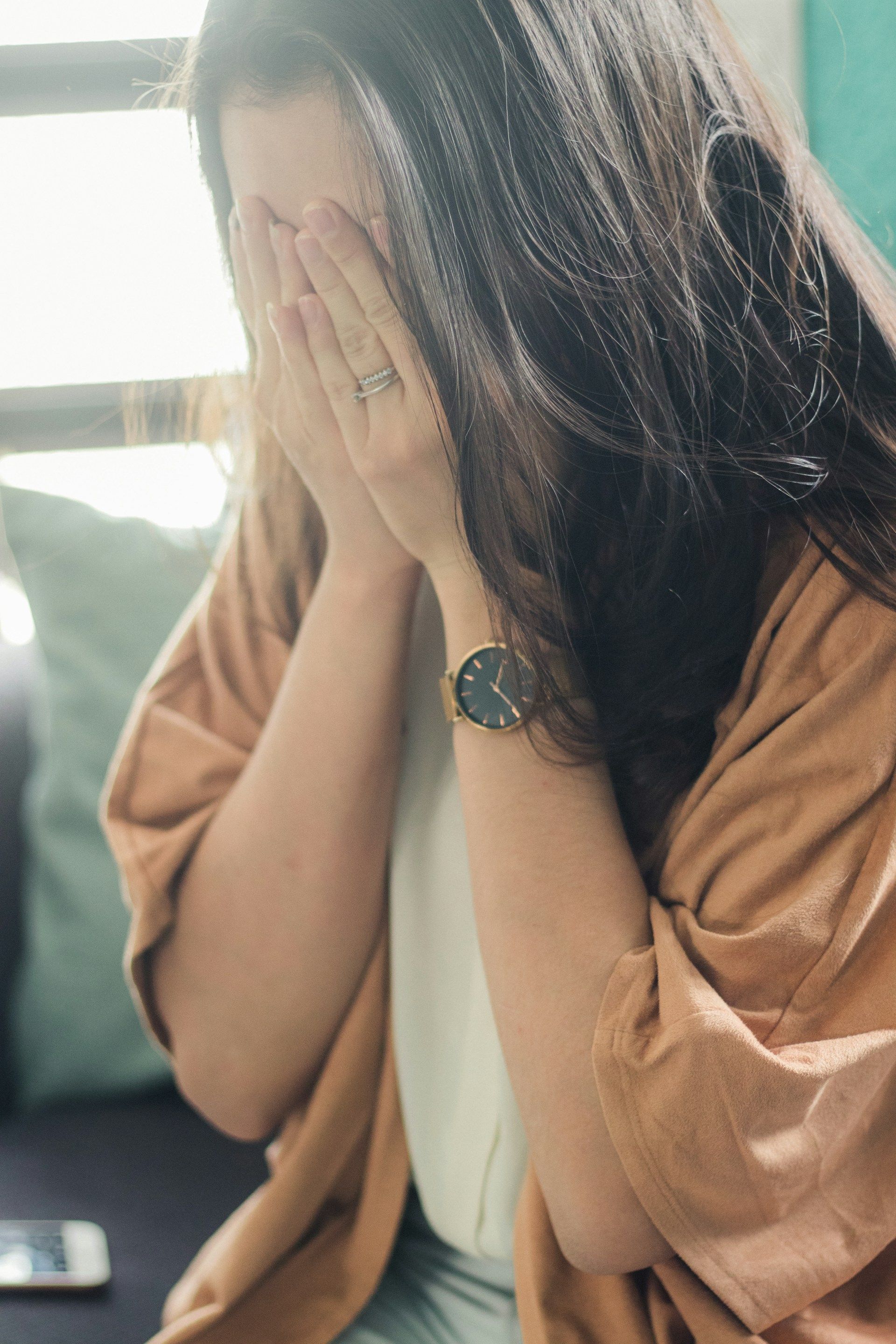 Woman with dark hair covering face, wearing a watch and ring, appears distraught.