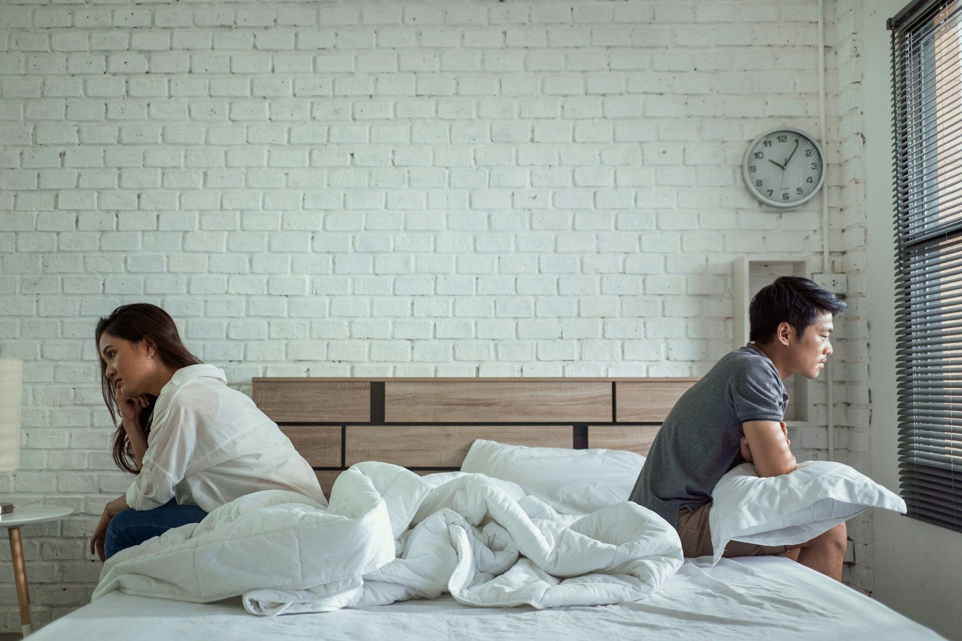 A couple sitting on a bed, back-to-back, looking sad. White bedding and brick wall.