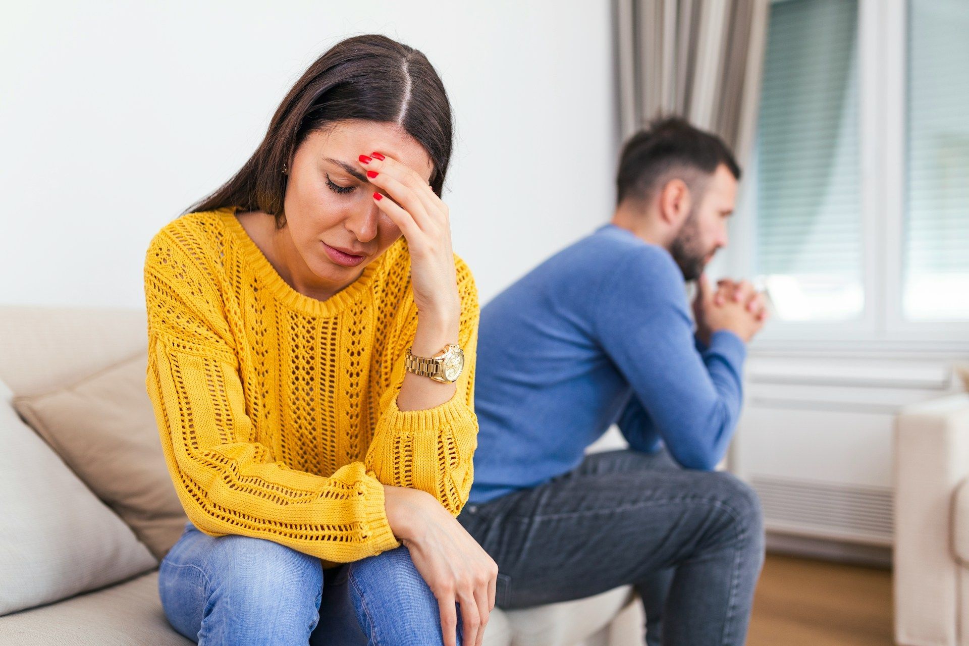 Woman in yellow sweater with head in hand, sits dejected next to a man looking away, indoors.