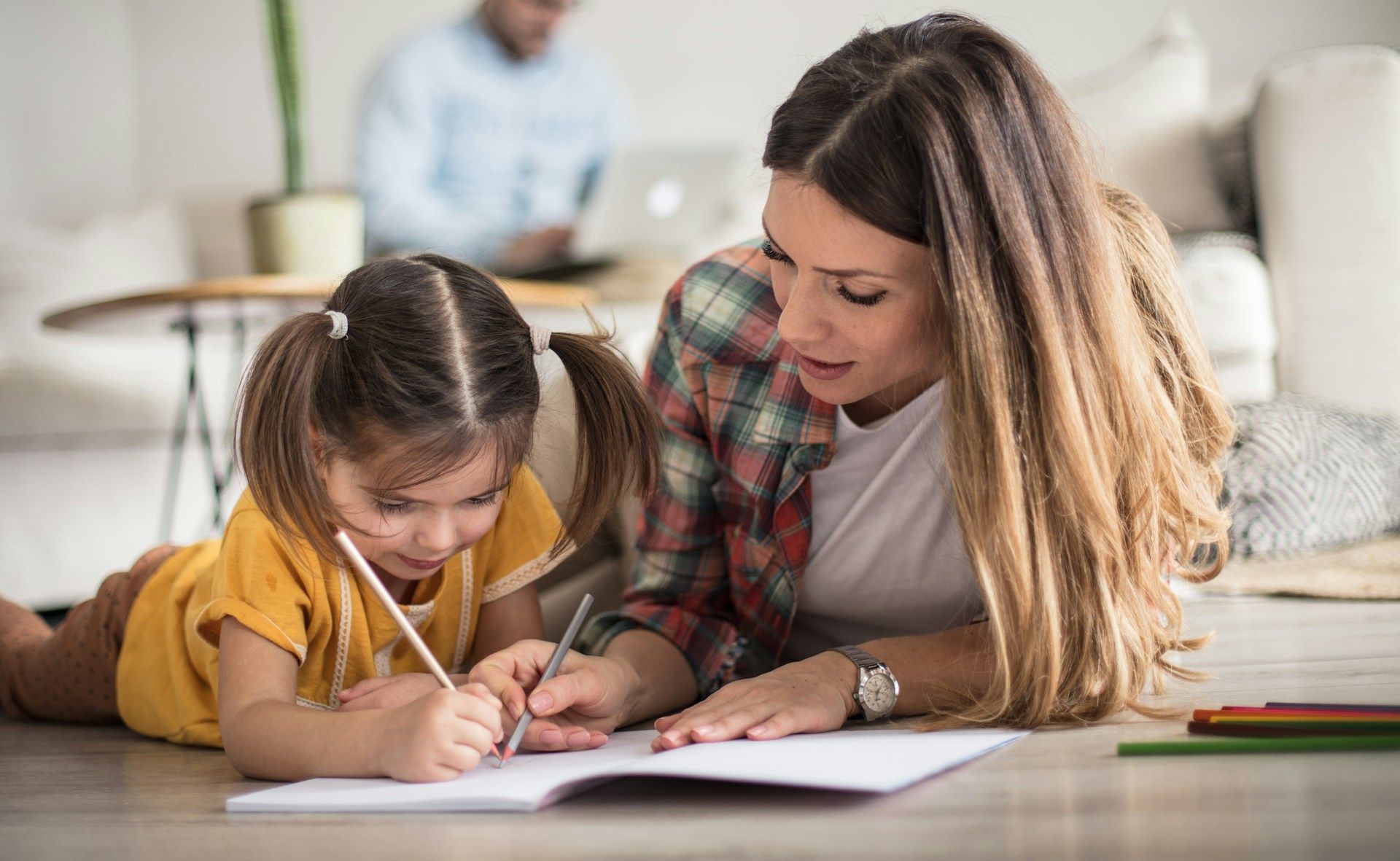 Woman and girl drawing together on the floor; man works on laptop in the background.