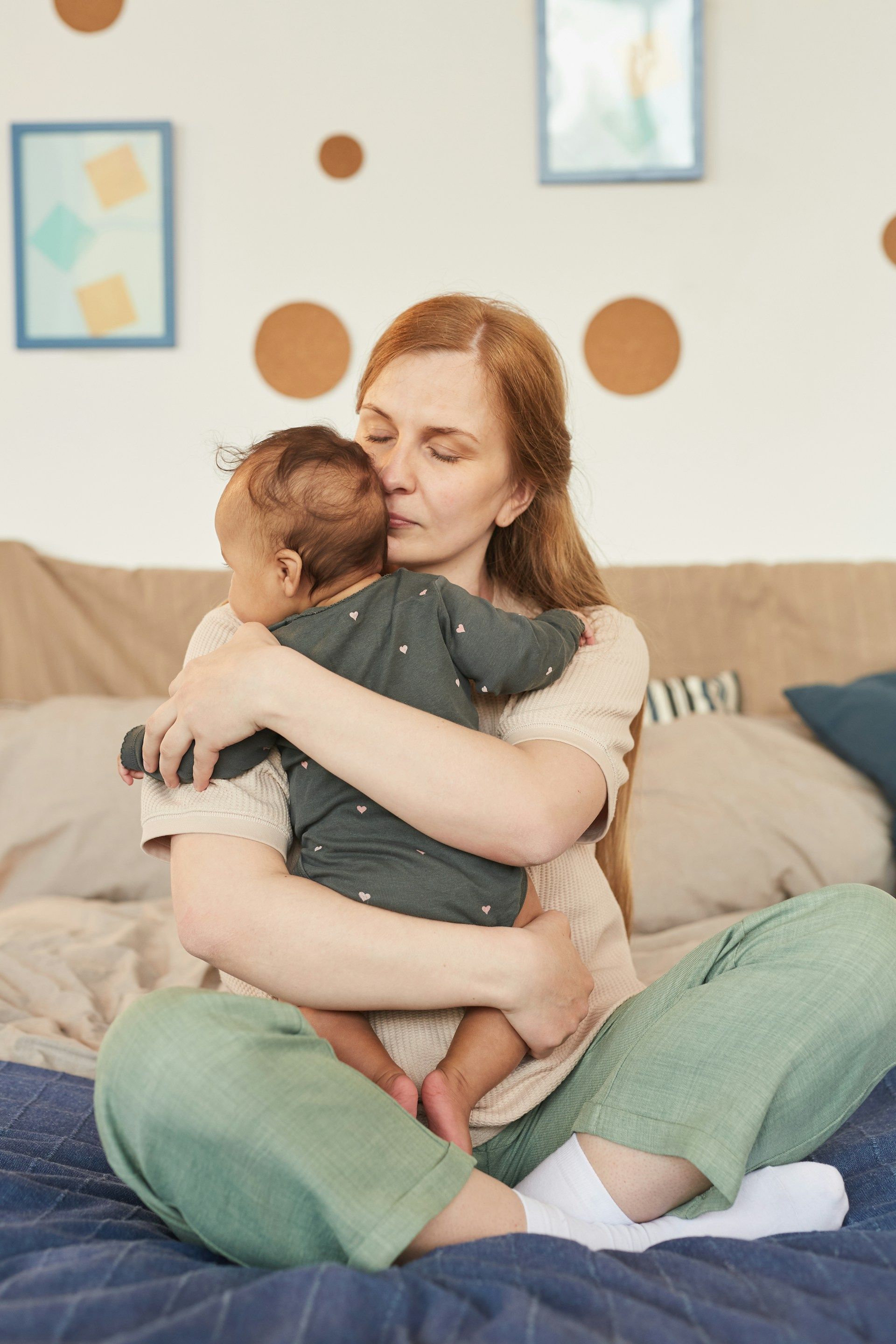 Woman in green pants sitting on bed, hugging and kissing a baby.