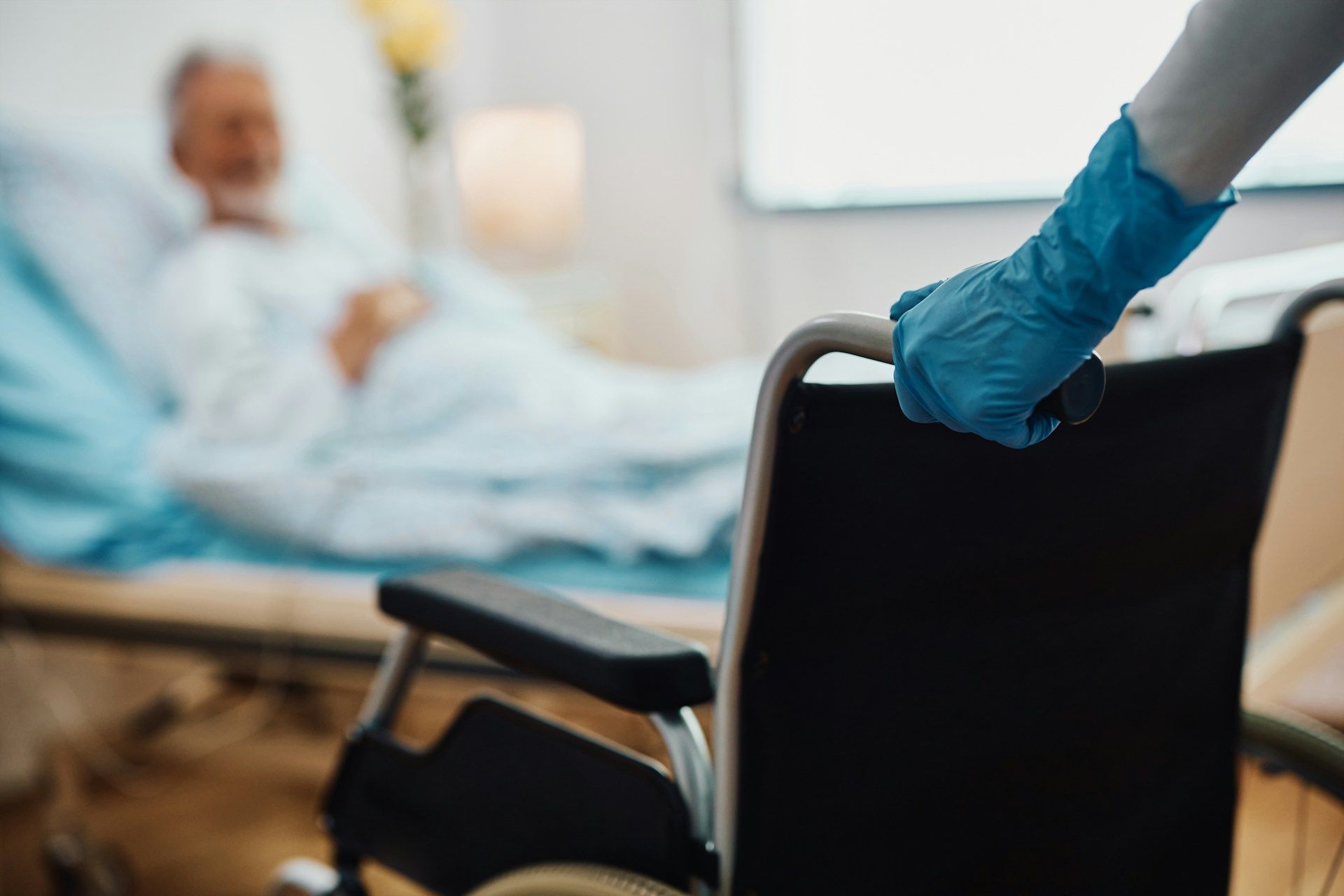 A person wearing a blue glove pushes a wheelchair in a hospital room, with a patient in bed.