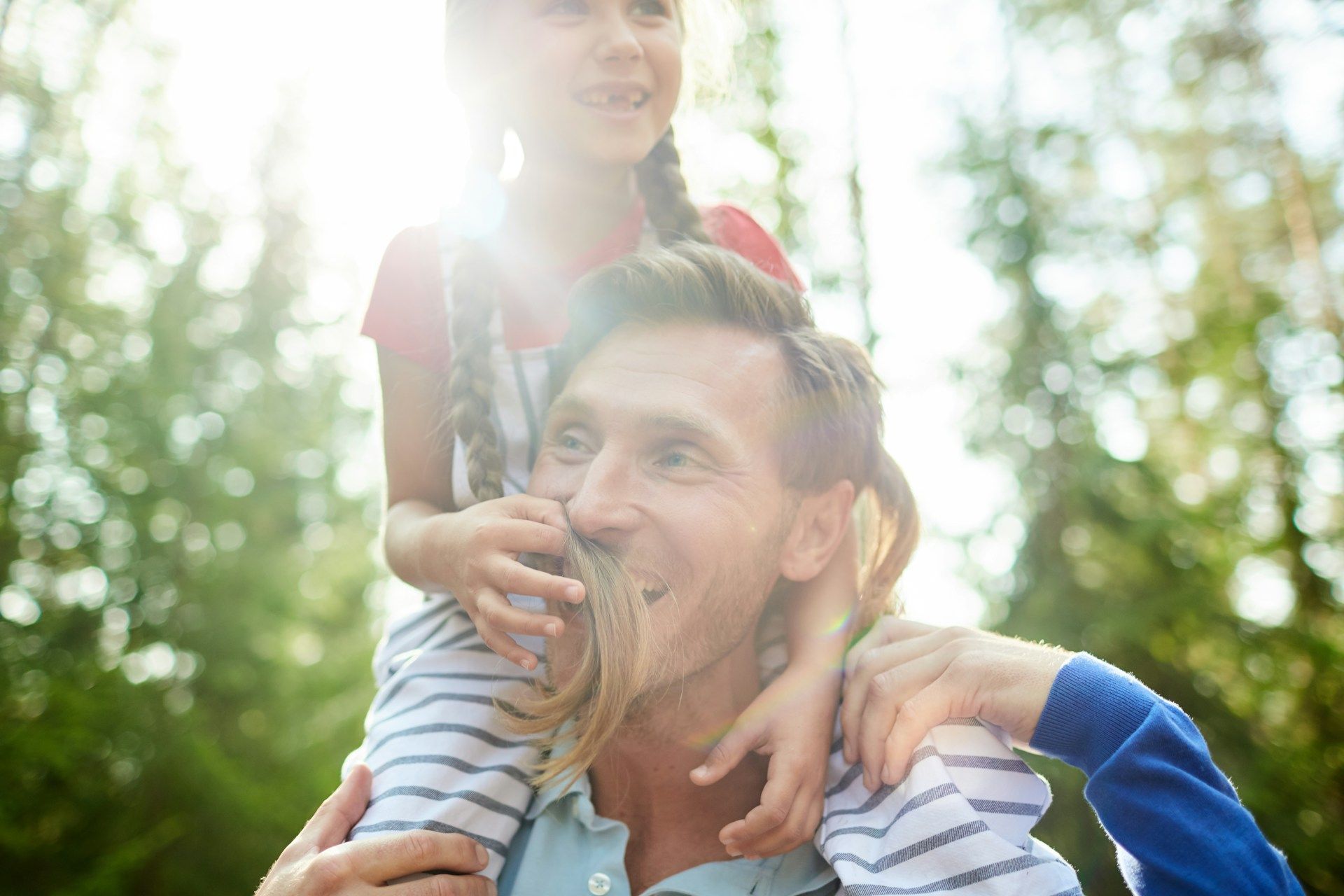 Man carrying a girl on his shoulders outdoors, smiling in sunlight.