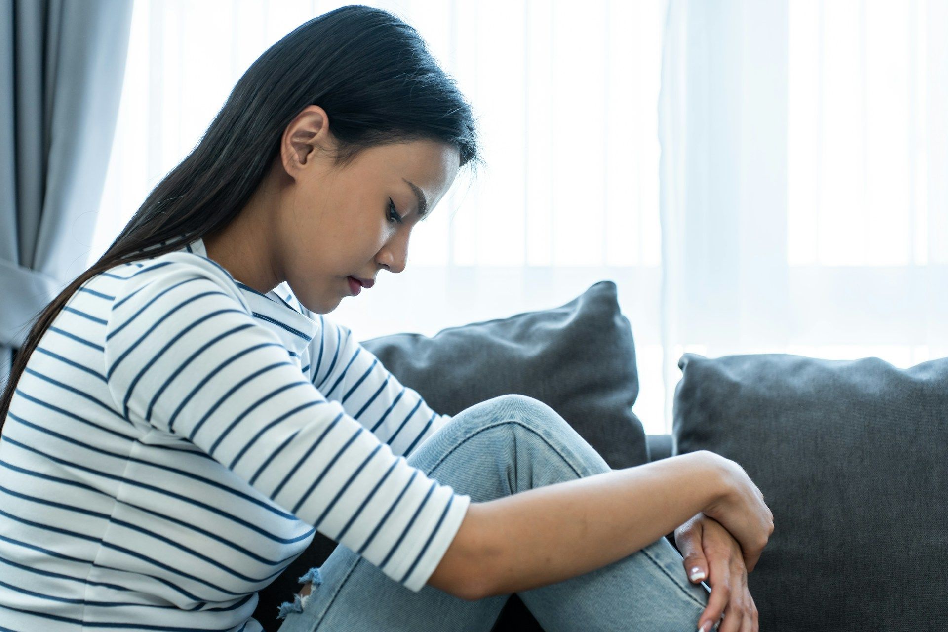 Woman sitting on a couch, looking down with a sad expression. Leans forward with arms wrapped around knees.